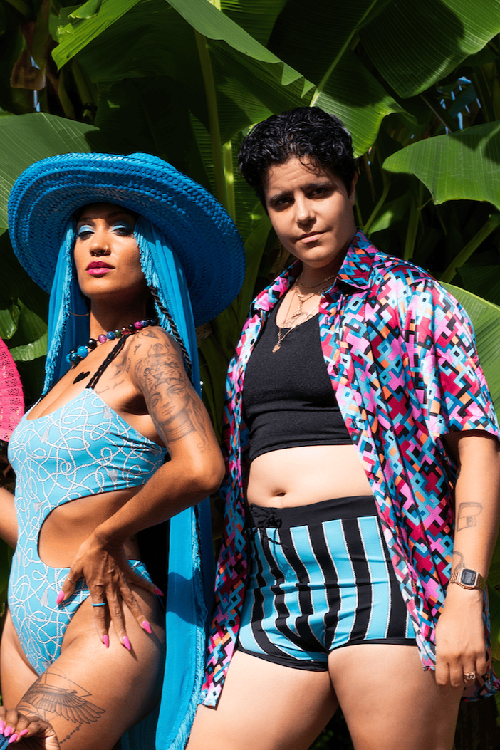 Two women in swimwear and cover-ups standing in front of large green leaves.
