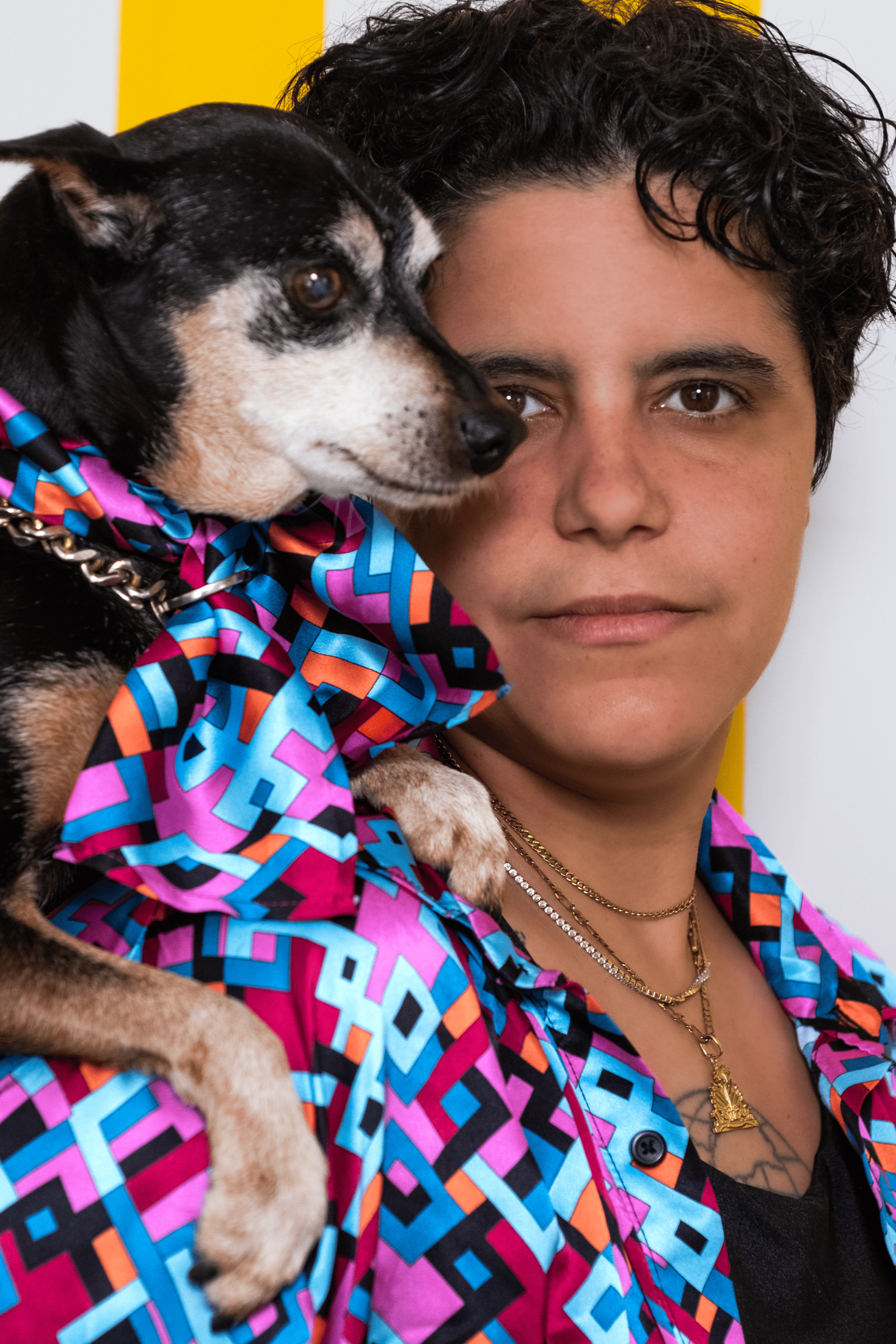 Person wearing a colorful jacket with a dog on their shoulder against a yellow and white striped background