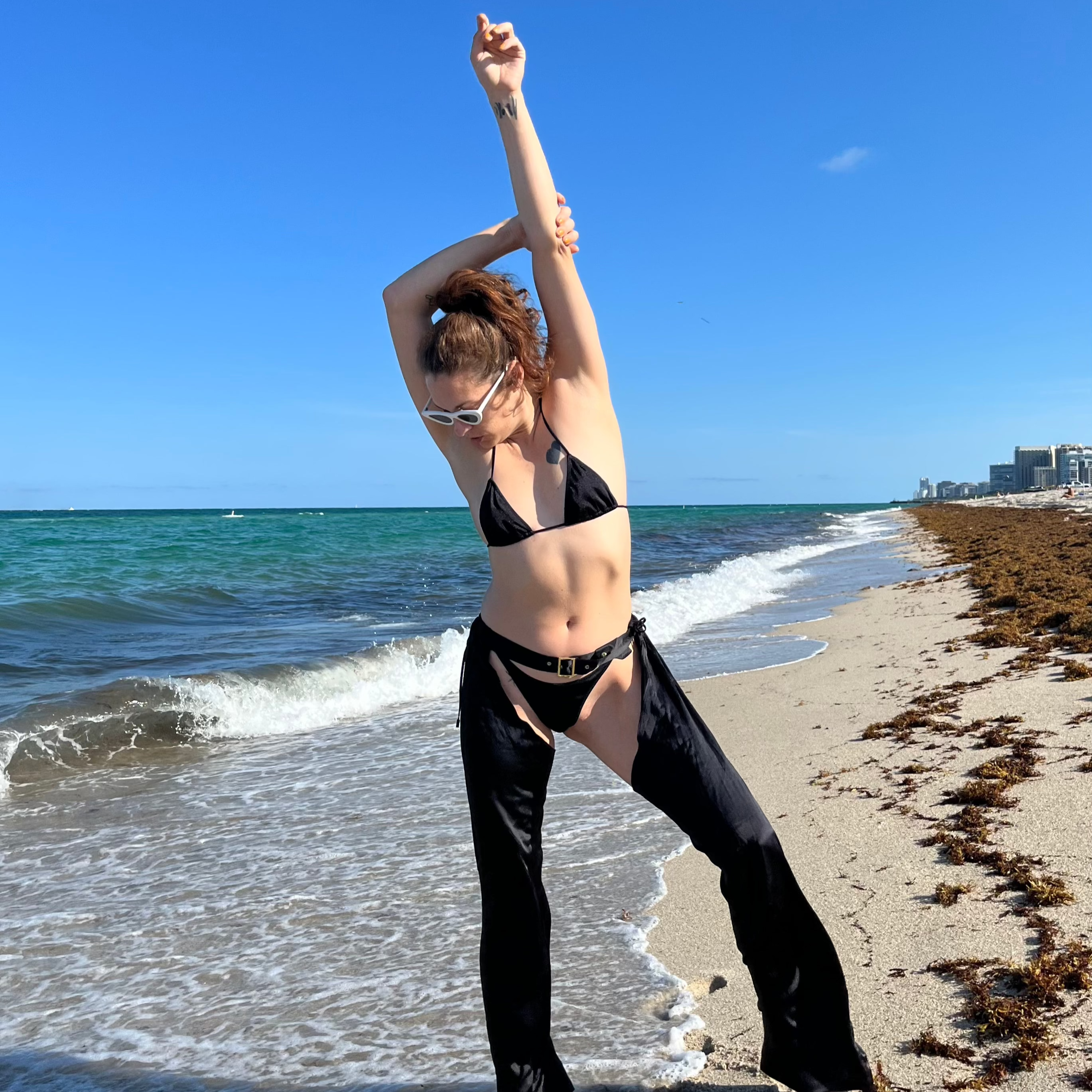Dani Read in a bikini top, bikini bottom and black chaps posing on a beach with ocean waves and clear sky.