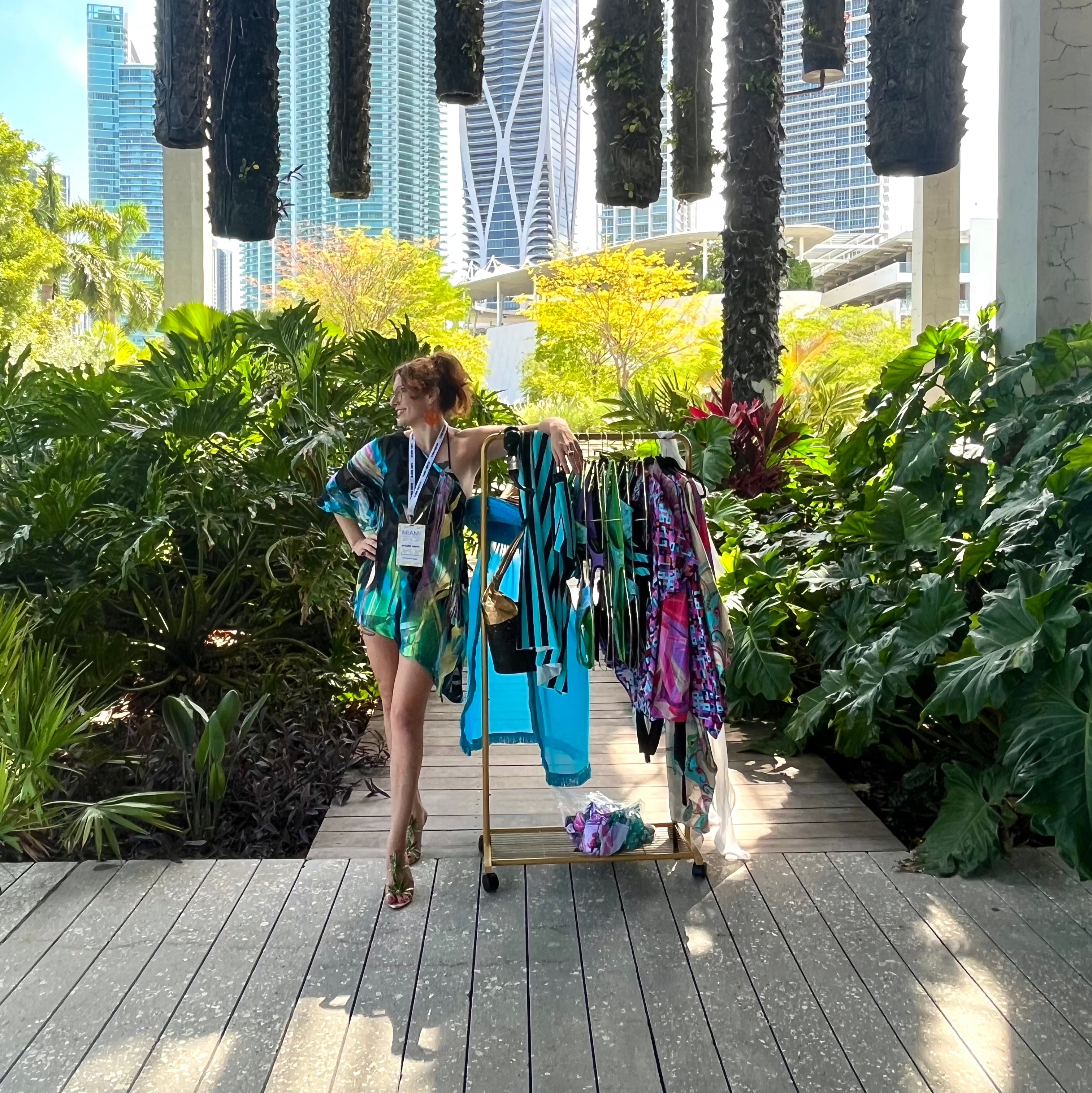 Dani Read with a rack of colorful clothing in an outdoor setting with plants and city skyline.